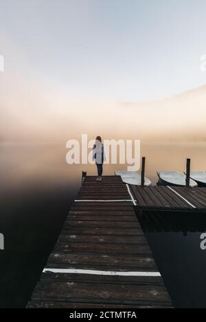 Junge Reisende Frau entspannen auf dem hölzernen Pier in der Nähe see, und beobachten Sie den nebligen Sonnenaufgang und genießen Sie die schöne Natur In der Wildnis für Stockfoto