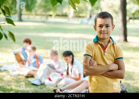 Portrait von lächelnd selbstbewussten Jungen in gelbem T-Shirt stehen mit gekreuzten Armen im Sommer Park, Schüler tun Aufgabe im Hintergrund Stockfoto