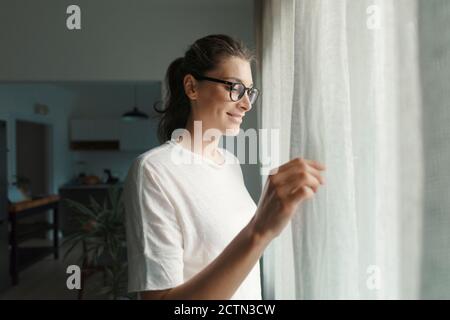 Glückliche Frau mit Brille neben einem Fenster stehen und öffnen Vorhänge zu Hause, Lifestyle-Konzept Stockfoto