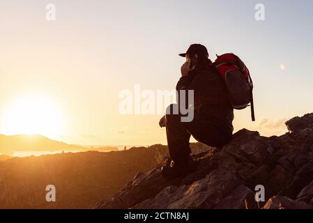 Seitenansicht eines nicht erkennbaren männlichen Wanderers, der auf einem Felsen sitzt highlands und sprechen am Handy, während Sie den Sonnenuntergang bewundern Stockfoto