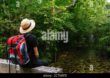 Entspannte weibliche Tourist mit Rucksack sitzt entlang Steinzaun und Wegschauen Stockfoto