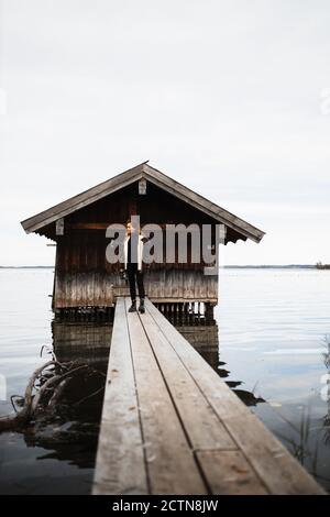 Nicht erkennbare weibliche Reisende in warmer Kleidung auf Holzbrett stehen pier in der Nähe von Shabby Shed am See im düsteren Herbst Tag Stockfoto