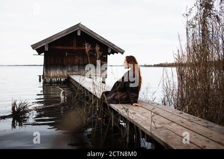 Weibliche Reisende in warmen Kleidern sitzen auf hölzernen Plank Pier In der Nähe schäbigen Schuppen am See in düsteren Herbsttag gelegen Stockfoto