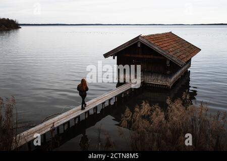 Nicht erkennbare weibliche Reisende in warmer Kleidung auf Holzbrett stehen pier in der Nähe von Shabby Shed am See im düsteren Herbst Tag Stockfoto