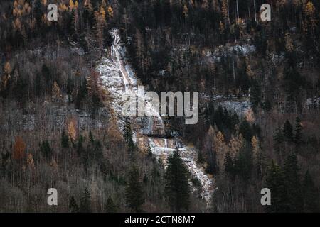 Erstaunliche Fernsicht auf gefrorenen verschneiten Wasserfall fließt auf den Berg Hang bedeckt mit Nadelwäldern in kalten Herbsttag Stockfoto