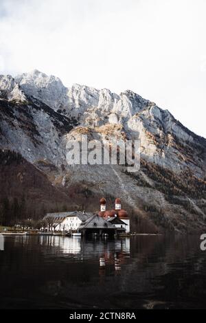 Malerische Landschaft mit kleinen Dorfhäusern und Kirche befindet sich auf Ufer des ruhigen Sees gegen raue felsigen Hang des Berges Bedeckt mit Schnee bei sonnigem Herbstwetter Stockfoto