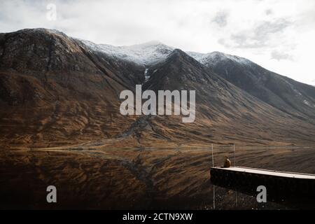 Fernsicht auf einsame Touristen sitzen auf hölzernen Pier und Bewundern Sie die friedliche Landschaft der Berge und See an bewölkten Tag In den schottischen Highlands Stockfoto