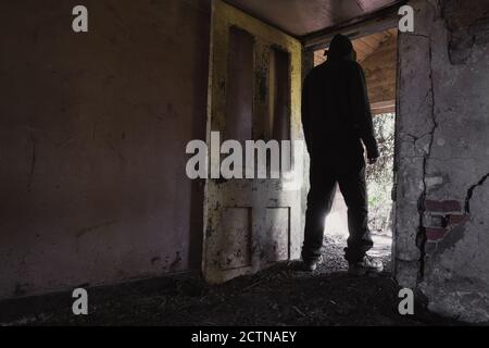 Ein Mann mit Kapuze, zurück zur Kamera, steht in der Tür einer zerstörten, verlassenen Hütte. Stockfoto