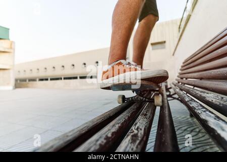 Niedriger Winkel der unkenntlichen Ernte männlich auf Skateboard auf Holzbank und zeigt Stunt Stockfoto