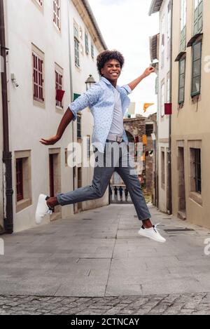 Seitenansicht des sorglosen afroamerikanischen Mannes im Moment der Über die Stadtstraße springen und die Kamera betrachten Stockfoto