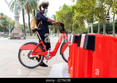 Junger schwarzer Mann in einem Anzug und Maske hören Musik mit dem öffentlichen Fahrrad auf der Straße Stockfoto