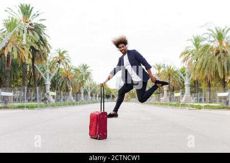 Junger schwarzer Mann in einem Anzug springen und hören Musik, während Sie einen Koffer auf Rädern umklammert und der Kamera zugewandt sind Auf der Straße Stockfoto