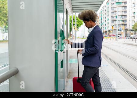 Schwarzer Mann mit Afro-Haaren zieht ein Ticket auf Die Straßenbahn mit einem Koffer neben ihm und ein Schnurloses Headset um den Hals Stockfoto