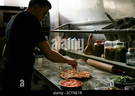 Seitenansicht des männlichen Pizzabackers, der frisch gebacken und appetitlich schneidet Heiße Pizza auf der Marmortheke während der Arbeit in der Restaurantküche Stockfoto