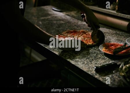 Crop anonymous Pizza Maker Schneiden frisch gebackene appetitlich heiße Pizza Auf Marmortisch während der Arbeit in der Restaurantküche Stockfoto