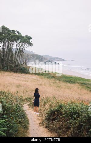 Rückansicht von unkenntlich einsamen Weibchen im schwarzen Kleid stehend Auf schmalem Weg, der durch grasbewachsenen Hügel zum Meer führt wolkiger Sommertag in Asturien in Spanien Stockfoto