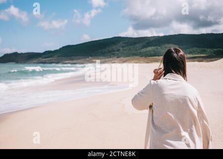 Rückansicht einer unerkennbaren Reisenden, die barfuß auf leer steht Sandstrand in der Nähe winken Meer und genießen frische Brise während Sommerurlaub in Spanien Stockfoto
