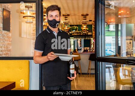 Seriöse Kellner stehen mit einer Flasche Bier und Teller hinein Cafe beim Betrahren der Kamera Stockfoto