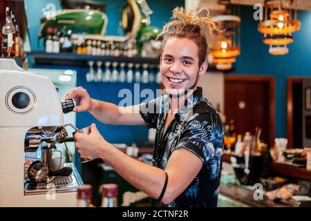 Seitenansicht des fröhlichen männlichen Barista mit Dreadlocks Vorbereitung aromatisch Kaffee in der Kaffeemaschine, während man die Kamera betrachtet