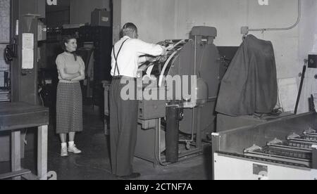 1940er Jahre, historisch, in einem Druckraum, eine Dame, die einem Ausflügler in Hosen und Hosenträger zusieht, und seine Jacke abschneidet, einen Streifen Papier auf einer Maschine ausschneidet, die Fotos von Filmnegativen, USA, druckte. Die Maschine war eine Kodak Papierverarbeitungsmaschine. Stockfoto