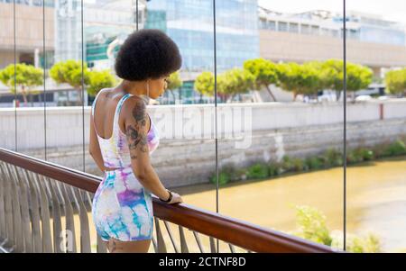 Seitenansicht einer entspannten afroamerikanischen Hündin, die auf der Aufhängung steht Brücke und bewundern Sie den Fluss in der Stadt während eines Spaziergangs An sonnigen Tag Stockfoto