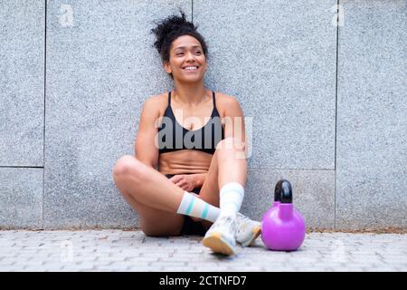 Positive schwarze athletische Frau in Sportkleidung sitzt auf Bürgersteig mit kettlebell und Pause beim Functional Workout in der Stadt Stockfoto