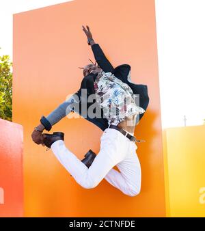 Niedriger Winkel von sorglosen afroamerikanischen Männchen in ausgefallenen Outfit Im Moment des Sprüngens über dem Boden in der Nähe von vibrierenden orange Wand In der Stadt Stockfoto