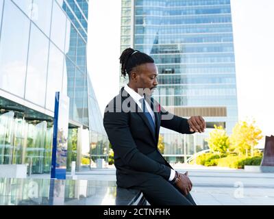 Seitenansicht des ernsten afroamerikanischen männlichen Unternehmers beim Betrachen Armbanduhr und Zeitkontrolle beim Sitzen in der Stadt und Warten Für Besprechungen Stockfoto