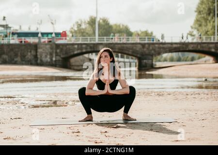 Konzentrierte Frau in Sportbekleidung Yoga in Malasana üben beim Balancieren Auf Matte am Sandstrand in der Nähe des Flusses, während Sie weg schauen Stockfoto