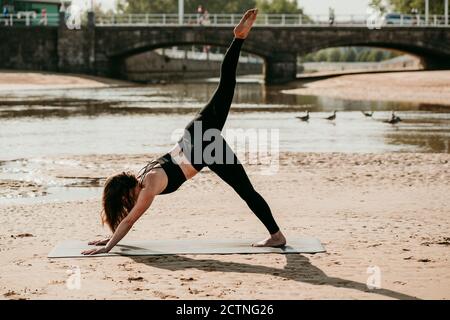 Seitenansicht einer schlanken Frau, die Yoga in Eka Pada praktiziert Adho Mukha Svanasana, während sie auf Matte am Flussufer in Sommer Stockfoto