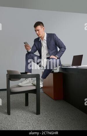 Beschäftigt männlichen Unternehmer sitzen auf Tisch mit Beinen auf Stuhl Am Arbeitsplatz und mit dem Smartphone, während Sie wegschauen Stockfoto