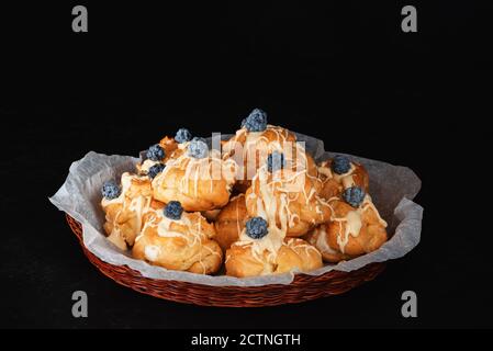 Französisches Dessert. Profiteroles mit Pudding Creme verziert mit geschmolzener weißer Schokolade und Dewbeeren in Korbkorb auf schwarzem Hintergrund. Low-Taste Stockfoto