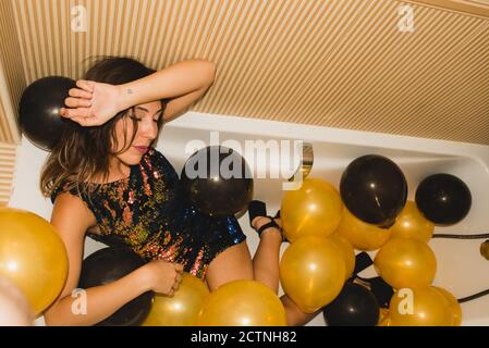 Von oben der jungen Frau im festlichen Kleid schlafen in Badewanne mit bunten Luftballons nach Feiertagsfeier gefüllt Stockfoto