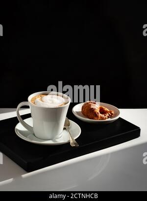 Serviert weiße Keramikbecher mit frischem Kaffee mit Milch gekrönt Schaum und Croissant auf dem Teller Stockfoto