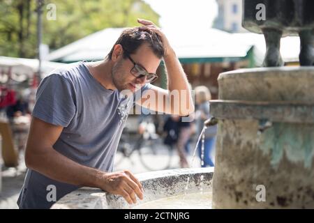 Junge Casual cucasian Mann erfrischt sich mit Wasser aus der Öffentlichkeit Stadtbrunnen an einem heißen Sommertag Stockfoto