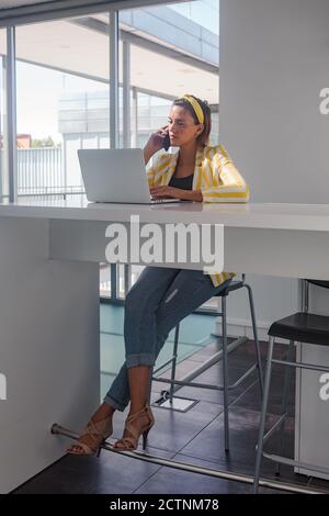 Moderne Freiberuflerin in stilvollem Outfit sitzt an der Theke mit Laptop und besprechen Details des Projekts über Handy während Arbeiten in modernen Arbeitsräumen Stockfoto
