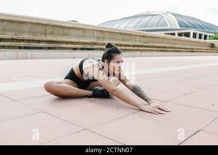 Ganzkörper der entschlossenen schlanken sportlichen Frau in activewear Durchführung Sitzende Vorwärtsbeugeübung und Stretching Körper während Fitness-Training Auf einer asphaltierten Straße Stockfoto