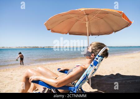 Frau trägt eine Gesichtsmaske im Sommerurlaub liegend in Die Sonne auf einer Liege direkt neben an Orangefarbener Regenschirm, während ihr Sohn am Ufer spielt TH Stockfoto