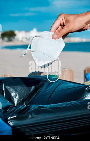 Nahaufnahme eines kaukasischen Mannes, der einen gebrauchten weißen Wegwerfartikel wirft Gesichtsmaske zum Müll neben dem Strand Stockfoto