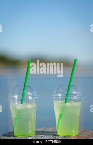 Medium shot of a glass filled to the top green drinking liquid, green straws with ice cubes with blurred blue ocean and sky in the distance Stockfoto