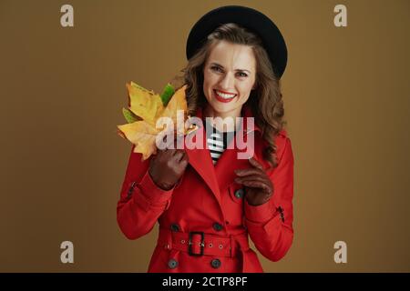 Hallo november. Portrait von lächelnder junger Frau in rotem Mantel und schwarzer Baskenmütze mit Lederhandschuhen und gelben Herbst Ahornblättern isoliert auf Bronze Bac Stockfoto