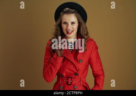 Hallo oktober. Glückliche moderne 40 Jahre alte Frau in rotem Mantel und schwarzer Baskenmütze mit Lederhandschuhen und Brille auf beigem Hintergrund. Stockfoto