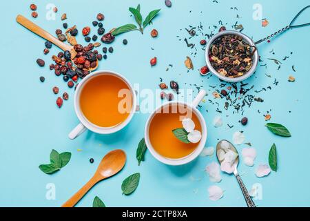 Kreativ arrangierte Tassen mit Tee auf einem klaren blauen Hintergrund. Isolierte Zusammensetzung mit Blütenblättern, grünen und getrockneten Blättern, Beeren. Stockfoto