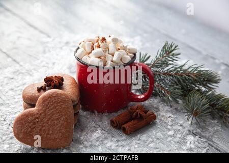 Tasse heißer Kakao mit Marshmallows und weihnachtlicher Lebkuchen in Form eines Herzens auf einem verschneiten Holzhintergrund. Weihnachtskarte. Stockfoto
