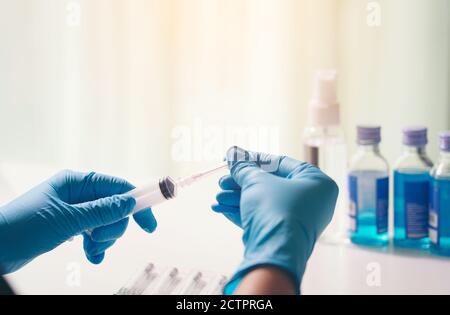 Nette Ärztin in einer medizinischen Schutzmaske hält eine Spritze mit einem Impfstoff in den Händen. Vor dem Hintergrund ist ein Mann in einer schützenden Chirurgie Stockfoto