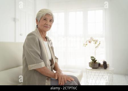 Lächelnde ältere Frau, die auf dem Sofa sitzt und die Kamera anschaut. Wecken Sie die alte Frau mit grauen Haaren und Schlafanzug im frühen Morgenlicht. Porträt von Elderl Stockfoto