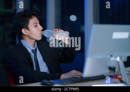 Junge asiatische gut aussehende Geschäftsmann Trinkwasser nach der Arbeit hart im Büro in der Nacht, Geschäftsleute entspannen nach der Arbeit, Geschäftserfolg Konzept Stockfoto