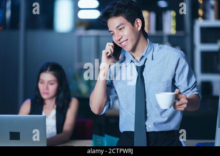 Geschäftsleute, die nachts im Büro arbeiten, Kaffee trinken und telefonieren, arbeiten zusammen über einen Laptop an einem Schreibtisch in einem Büro in La Stockfoto