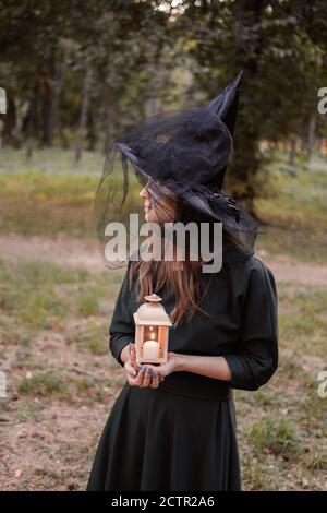 Junge Frau in dunklem Kleid und Hexenhut hält Laterne mit Kerzen in den Händen und beleuchtet den Wald. Halloween Party Kostüm. Parken mit Autum Stockfoto