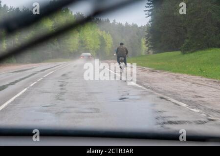 Blick durch die Windschutzscheibe in Regentropfen: Ein Mann fährt auf der Straße mit dem Fahrrad, ein schwarzes Auto fährt an seiner Seite vorbei Stockfoto